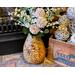 A handmade sunflower vase,  filled with faux daisies and zinnias, decorates a fireplace hearth. The vase is made from a dried gourd. 